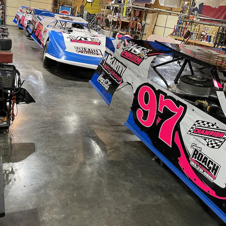 Dirt track race cars with a pink, blue and white wrap design, number 97, parked in a garage